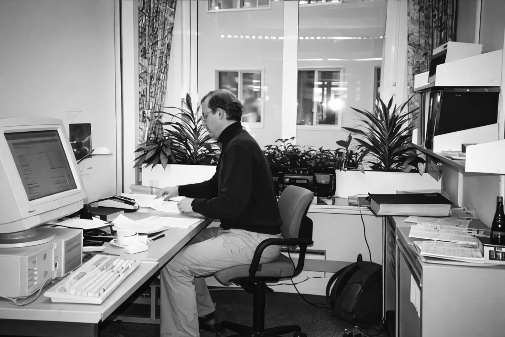 Czarno-białe zdjęcie biura z lat 90., pracownik przy biurku z komputerem CRT, za nim rośliny doniczkowe na parapecie i naturalne światło z okna.